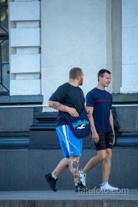 Фотография двух мужчин с татуировками на руках и ногах, идущих по улице. Видны детализированные рисунки татуировок в различных стилях.