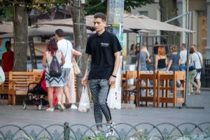 Фотография мужчины, идущего по улице с видимой татуировкой на предплечье. Он одет в черную футболку и серые брюки.