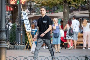 Фотография мужчины, идущего по улице с видимой татуировкой на предплечье. Он одет в черную футболку и серые брюки.