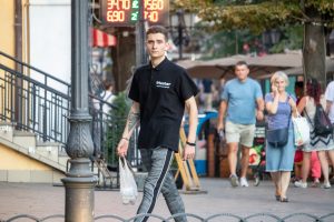 Фотография мужчины, идущего по улице с видимой татуировкой на предплечье. Он одет в черную футболку и серые брюки.