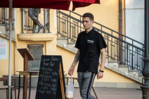 Фотография мужчины, идущего по улице с видимой татуировкой на предплечье. Он одет в черную футболку и серые брюки, держит пакет.