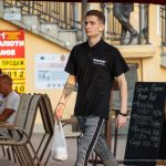 Фотография мужчины, идущего по улице с видимой татуировкой на предплечье. Он одет в черную футболку и серые брюки, держит пакет.