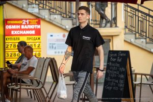 Фотография мужчины, идущего по улице с видимой татуировкой на предплечье. Он одет в черную футболку и серые брюки, держит пакет.