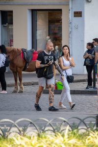 Фотография мужчины с татуировками на руках и шее, идущего по улице с девушкой. Татуировки выполнены в черном цвете, с использованием графических узоров и портретов.