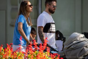 Фото мужчины с татуировкой на руке в стиле геометрии, женщины и ребенка. Темные узоры из шестиугольников покрывают предплечье. Семья гуляет на улице.