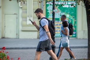 Фото мужчины с татуировкой на руке, идущего по улице и смотрящего в телефон. Современный городской стиль и минималистичный рисунок татуировки.
