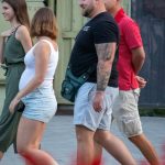 Мужчина с татуировкой на руке стоит рядом с беременной женщиной. Мужчина одет в черную футболку и шорты.