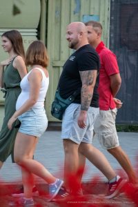 Мужчина с татуировкой на руке стоит рядом с беременной женщиной. Мужчина одет в черную футболку и шорты.