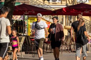 Фото тату: группа людей на улице, у одного из мужчин татуировка на руке. Городской пейзаж с кафе и магазинами.