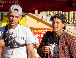 Фото тату: два мужчины на улице, один из которых с татуировками на руках и шее. Городской пейзаж с кафе и магазинами.