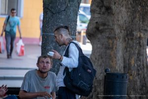 Мужчина с выбритыми волосами и татуировкой на шее разговаривает с другим человеком. На мужчине рваный рюкзак.
