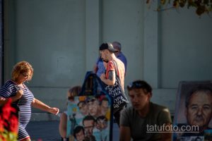 Фото тату на руке мужчины в кепке и футболке с принтом. Татуировка выполнена в черных тонах и видна на фоне ярких цветов.