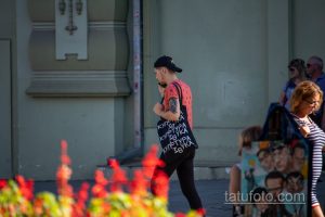 Фото тату на руке мужчины в кепке и футболке с принтом. Татуировка выполнена в черных тонах и видна на фоне ярких цветов.