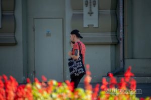 Фото тату на руке мужчины в кепке и футболке с принтом. Татуировка выполнена в черных тонах и видна на фоне красных цветов.