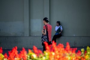Фото тату на руке мужчины в кепке и яркой футболке. Татуировка выполнена в черных тонах и видна на фоне красных цветов.
