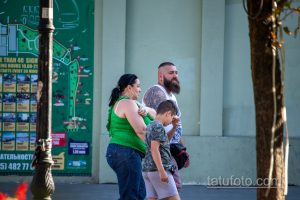 Фото тату на плече мужчины с бородой, гуляющего с семьей на улице. Татуировка выполнена в темных тонах и частично видна под одеждой.