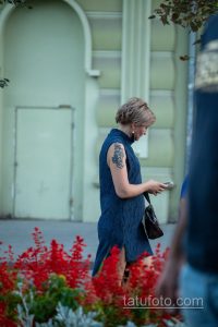 Фото татуировки на руке женщины: темный орнамент, напоминающий восточные мотивы, расположен на плече. Женщина смотрит в телефон.