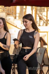 Фотография девушки с татуировкой цветка на плече и надписью 'freak mode' на майке. Девушка смотрит в телефон.
