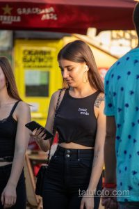 Фотография татуировки на плече девушки: реалистичный рисунок цветка с листьями, выполненный в черно-серых тонах. Девушка смотрит в сторону.