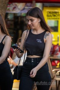 Фото татуировки на плече девушки: реалистичный рисунок цветка с листьями, выполненный в черно-серых тонах. Девушка смотрит в телефон.