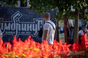 Фото тату на плече мужчины: крупный план полинезийской графики. Мужчина стоит на фоне ярких цветов и городской архитектуры.