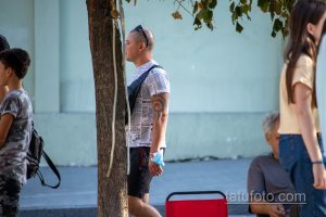 Фото татуировки на плече мужчины. Темный узор, напоминающий морской узел или якорь, на фоне газетных вырезок. Расположена на плече.