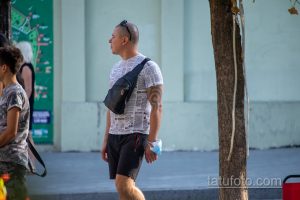 Фото татуировки на плече мужчины. Темный узор, напоминающий морской узел или якорь, на фоне газетных вырезок. Расположена на плече.