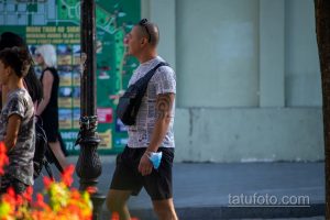 Фото татуировки на плече мужчины. Темный узор, напоминающий морской узел или якорь, на фоне газетных вырезок. Расположена на плече.