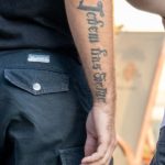 Фото тату: черная надпись на латыни, выполненная каллиграфическим шрифтом, расположена вдоль предплечья мужчины. Изящный рисунок татуировки.