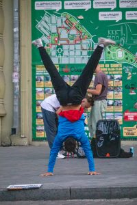 Фотография уличного акробата в динамичной позе, демонстрирующего татуировку на руке. Задний фон - городская стена с картой.