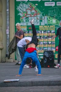Фотография уличного акробата, выполняющего стойку на руках на фоне городской стены с картой. Видна татуировка на спине акробата.