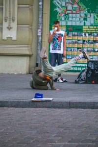 Фотография уличного акробата, выполняющего кувырок. На заднем плане видна карта города и мужчина с татуировкой на плече.