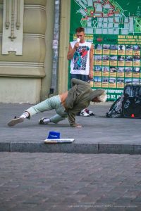 Фотография уличного акробата в движении, демонстрирующего гибкость и силу. На заднем плане видна карта города и мужчина с татуировкой на плече.
