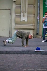 Фотография уличного акробата в движении, демонстрирующего гибкость и силу. На заднем плане видна архитектура города и рекламные плакаты.