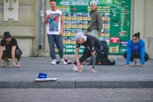 Фотография уличного акробата, выполняющего стойку на руках возле карты города. Видны татуировки на руках и плечах участников.