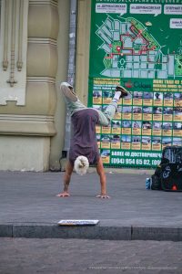 Фотография уличного акробата, выполняющего стойку на руках на фоне городской стены с афишами в Одессе. На руке видна татуировка.