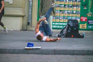 Фотография уличного акробата, выполняющего элемент. На руке акробата видна татуировка. Городской фон с афишами.