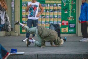 Фотография уличного акробата, выполняющего элемент. На руке акробата видна татуировка. Городской фон с афишами.