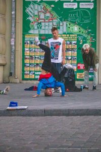 Фотография уличного акробата, выполняющего стойку на руках. На руке акробата видна яркая татуировка. Городской фон с афишами.
