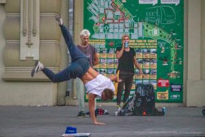Фотография уличного акробата, выполняющего сложный элемент. Видны татуировки на руках и плечах акробата на фоне городских афиш.