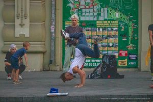 Фотография уличного акробата, выполняющего сложный элемент. Видны татуировки на руках и плечах акробата на фоне городских афиш.