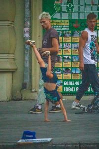 Фотография уличного акробата, выполняющего стойку на руках на фоне ярких афиш. Видны татуировки на руках и плечах акробата.