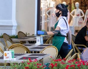 Фотография официантки в медицинской маске и голубых перчатках, выносящей заказы на террасе кафе. На заднем плане видны столики и витрины магазинов.
