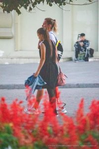 Фотография двух девушек, идущих по улице. Одна девушка обнимает другую, а на заднем плане видны цветы.