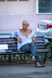 Фотография пожилого мужчины, сидящего на скамейке в городском парке. Мужчина выглядит задумчивым и уставшим.