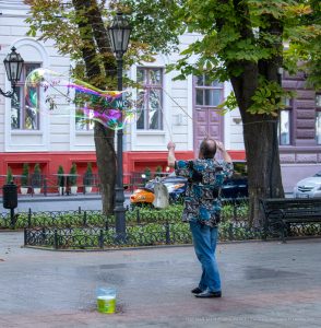 Фотография уличного артиста, создающего огромные мыльные пузыри на фоне городской архитектуры и деревьев.
