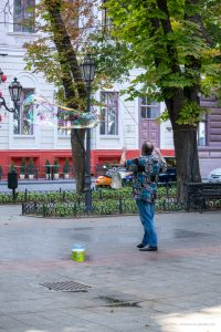 Фотография уличного артиста, создающего мыльные пузыри. Видны яркие пузыри, деревья и городская архитектура.