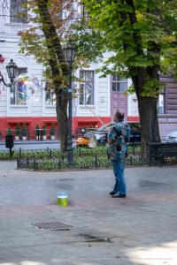 Фотография уличного артиста, создающего мыльные пузыри на фоне городской архитектуры и зелени. Видны деревья, фонари и прохожие.