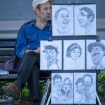 Фотография художника, рисующего карикатуры на улице. Рядом с ним развешаны готовые работы. Яркий солнечный день, городская сцена.