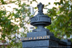 Фотография кованого элемента с надписью на постаменте. Фон размыт, видны зеленые листья.
